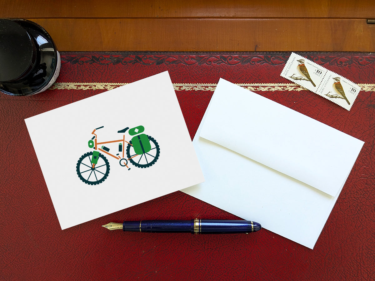 Greeting card with an orange bike with green travel bags on a table with a blue pen and envelope and stamps