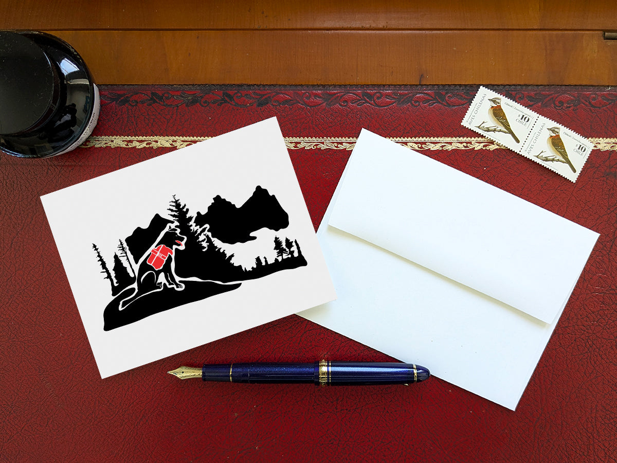 Greeting card with black dog with red backpack in the mountains on a table with blue pen and envelope and stamps.