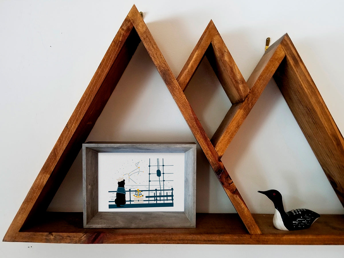 Framed art print with illustration of a dog looking at a constellation from a ship with a sextant on a wooden shelf.