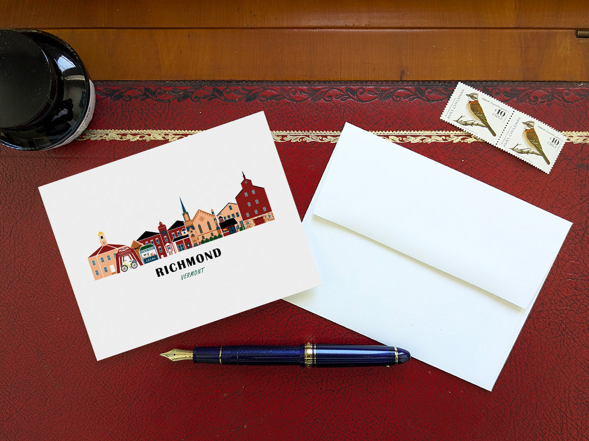 Greeting card with image of buildings arranged in a city skyline with the text Richmond Vermont on a table with blue pen and envelope and stamps.