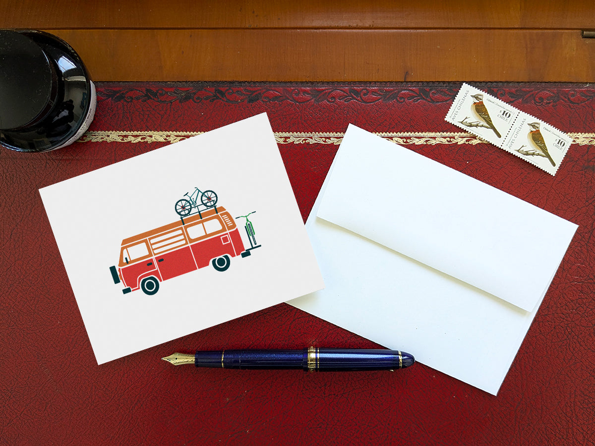 Greeting card with a red and orange camper van carrying two bikes on a table with a blue pen and envelope and stamps