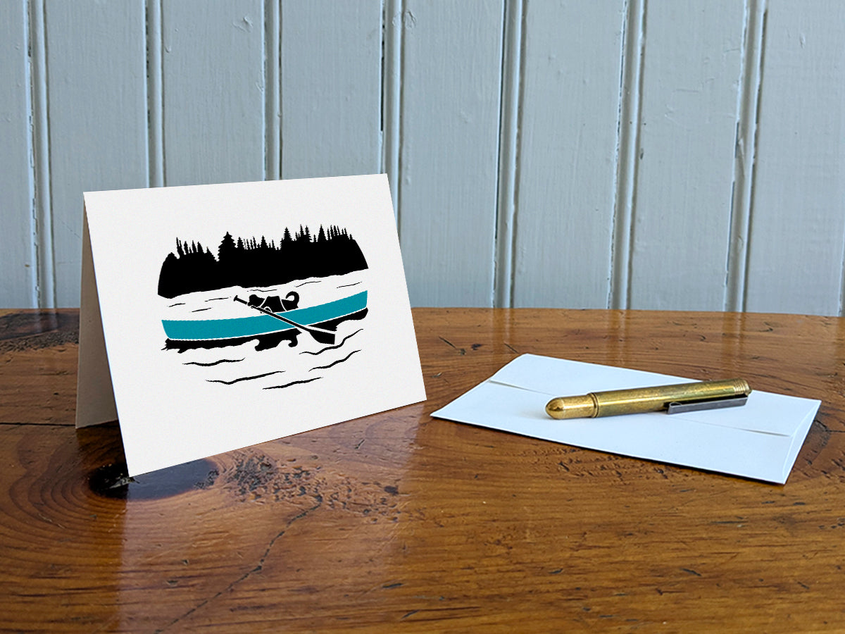 Greeting card with a black dog paddling a blue canoe on a table with a gold pen and envelope