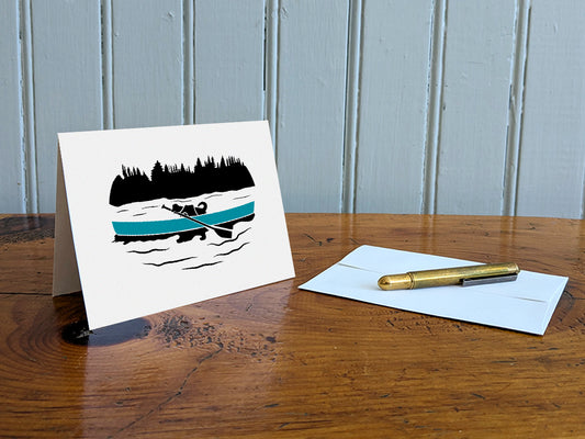 Greeting card with a black dog paddling a blue canoe on a table with a gold pen and envelope