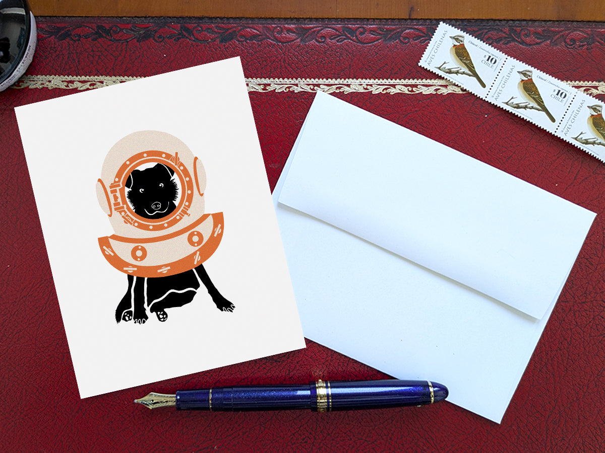 Greeting card with image of a dog with a scuba diving helmet on a table with a blue pen and envelope and stamps.