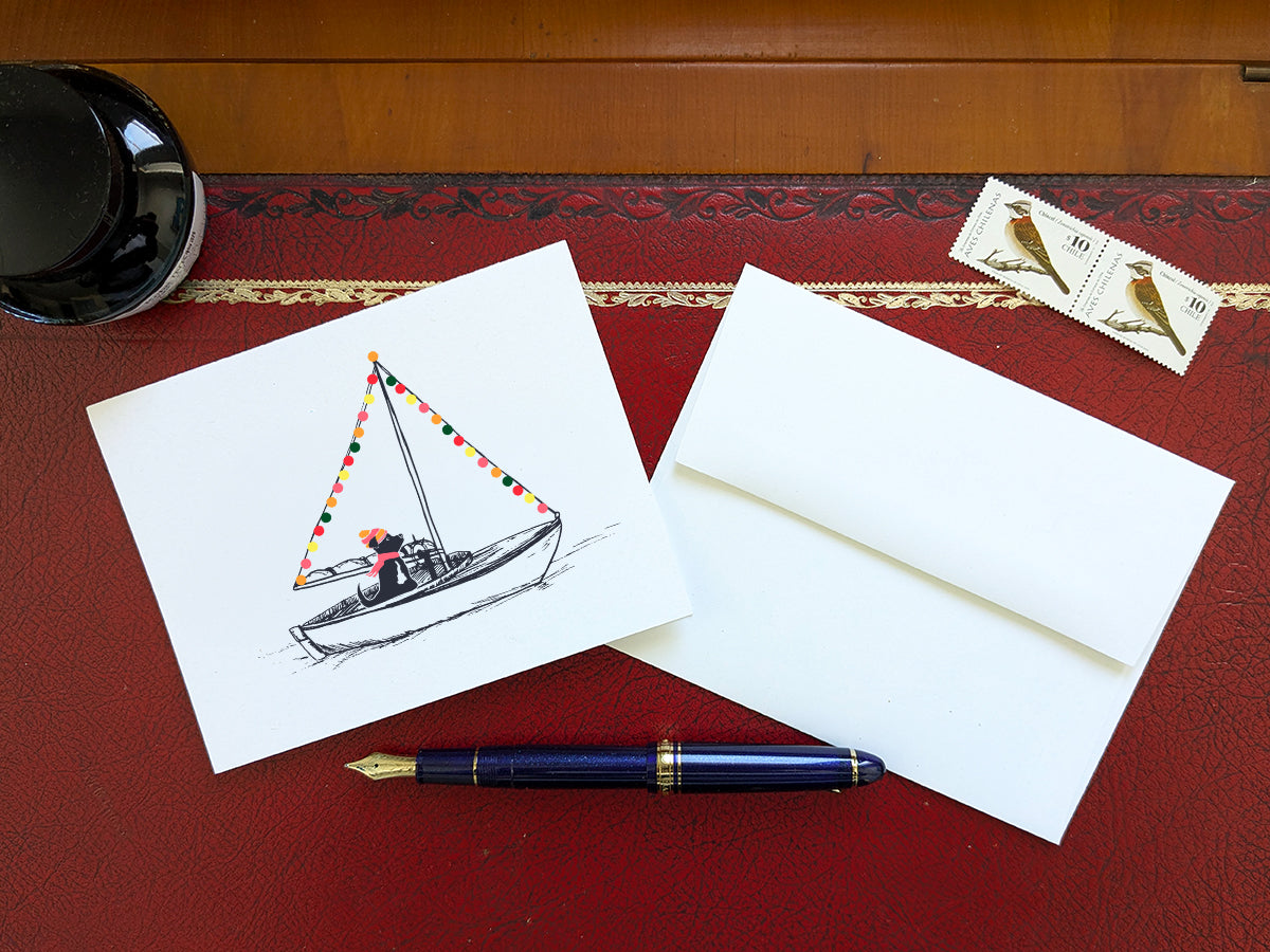 Greeting card with illustration of a black dog in a sailboat decorated with colorful lights, displayed on a red desk with a white envelope, stamps and blue fountain pen.