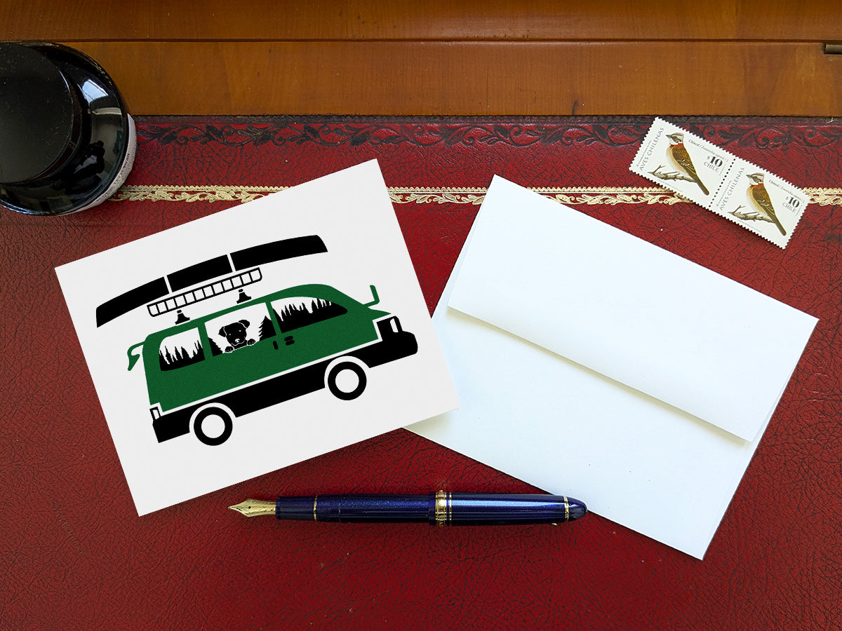 Greeting card with image of dog riding in a camper van with canoe on top on a table with blue pen and envelope and stamps.