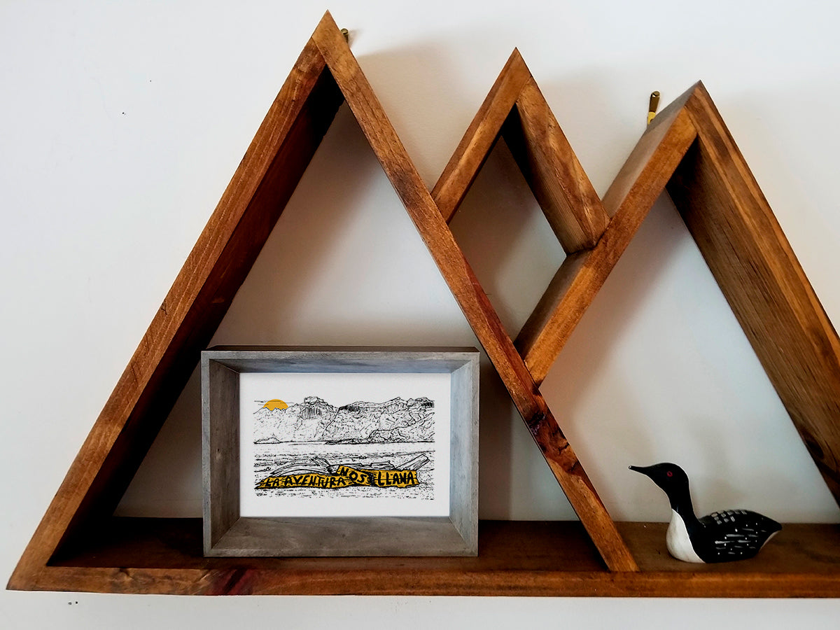 Framed art print on a wooden shelf of mountains and sunset with the text La aventura nos llama