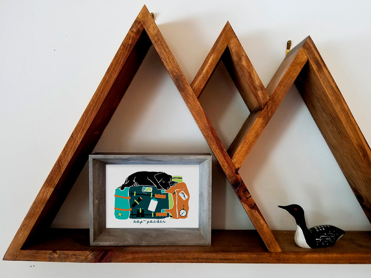 Framed art print on a wooden shelf of a black dog sleeping on top of a colorful backpack with the text nap-packer