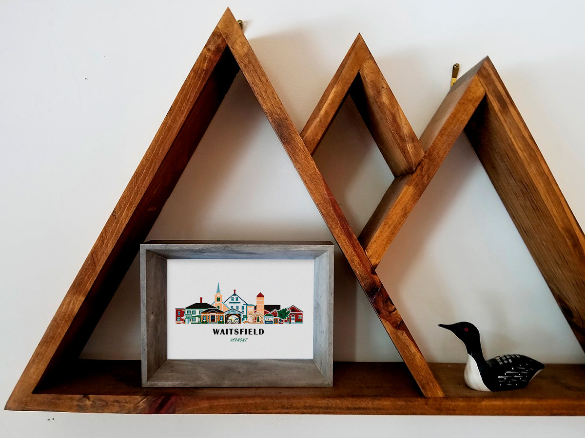 Framed art print on a wooden shelf of buildings arranged in a skyline with the text Waitsfield Vermont