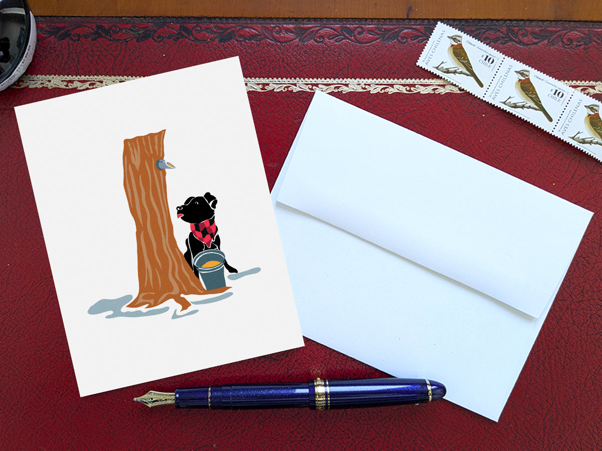 Greeting card with image of dog next to a maple syrup tap on a tree and bucket of maple syrup on a table with blue pen and envelope and stamps.