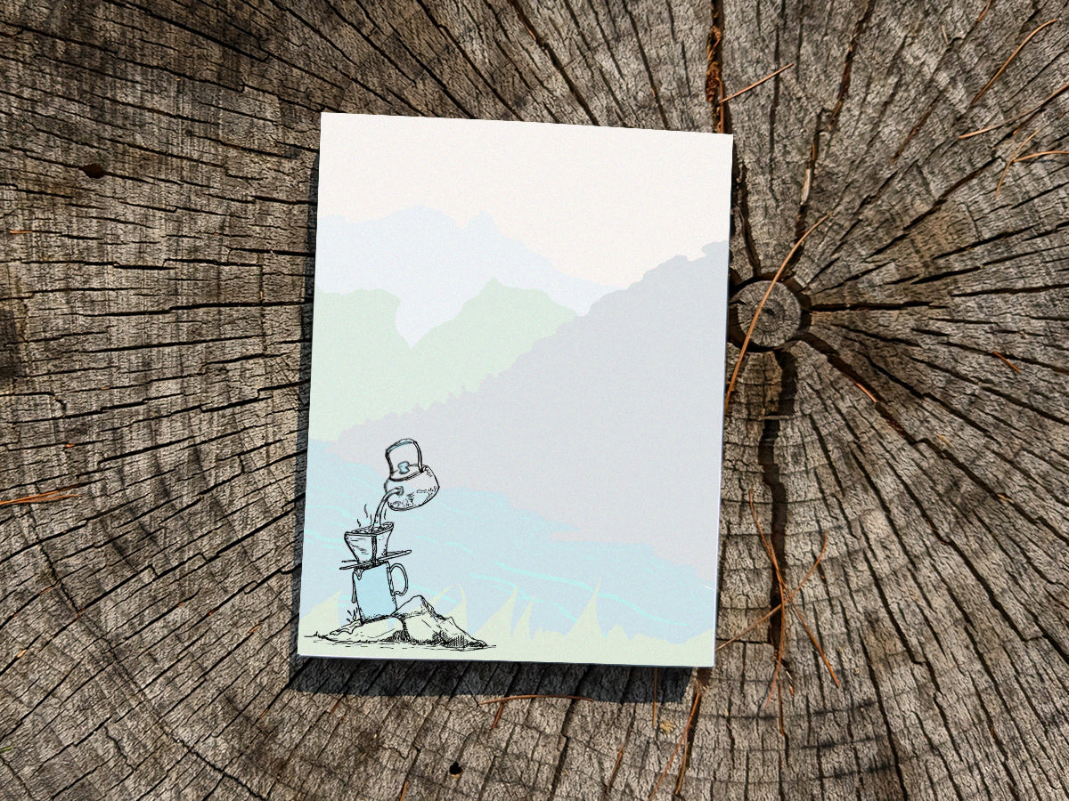 Notepad with illustration of pour-over coffee on a riverbank on a wood surface.