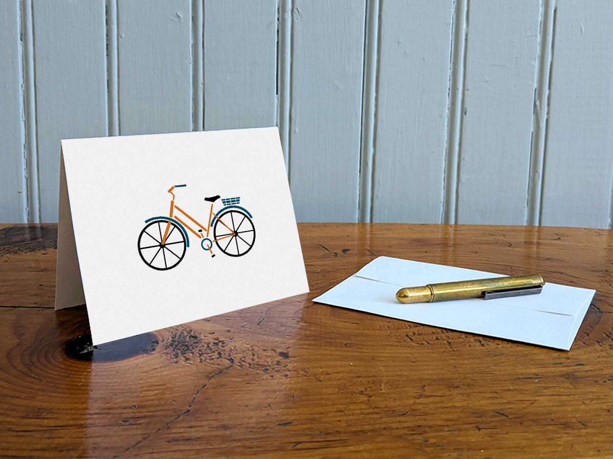 Greeting card with a yellow cruiser bike with basket on a table with gold pen and envelope