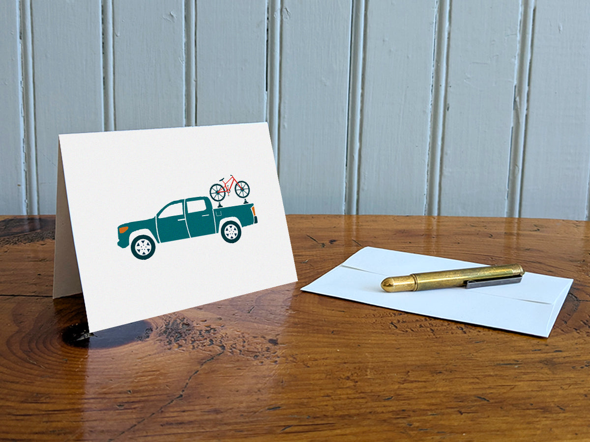 Greeting card of a blue truck carrying a bike on a table with gold pen and envelope