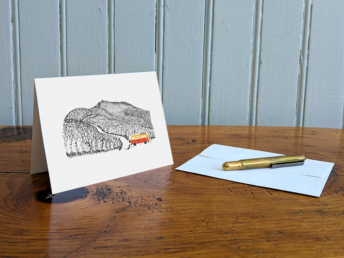 Greeting card with image of a mountain road and an orange and yellow campervan on a table with a gold pen and envelope.