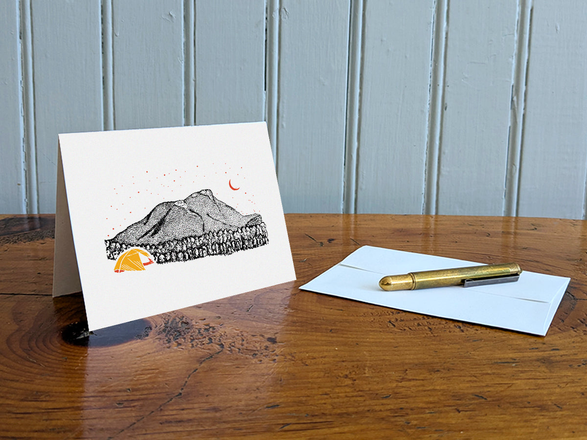 Greeting card with an image of a mountain and bright yellow tent under a starry sky on a table with gold pen and envelope.