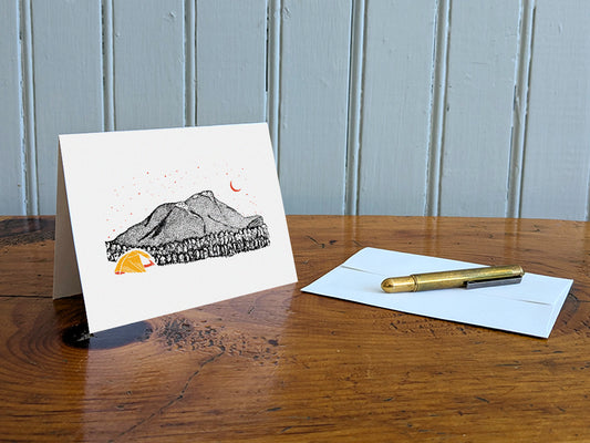 Greeting card with an image of a mountain and bright yellow tent under a starry sky on a table with gold pen and envelope.