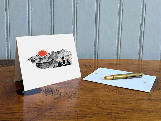 Greeting card with image of black dog looking at a red sunset behind mountains on a table with gold pen and envelope.