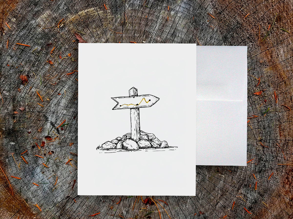White card with a black ink drawing of a trail marker on a wooden surface.
