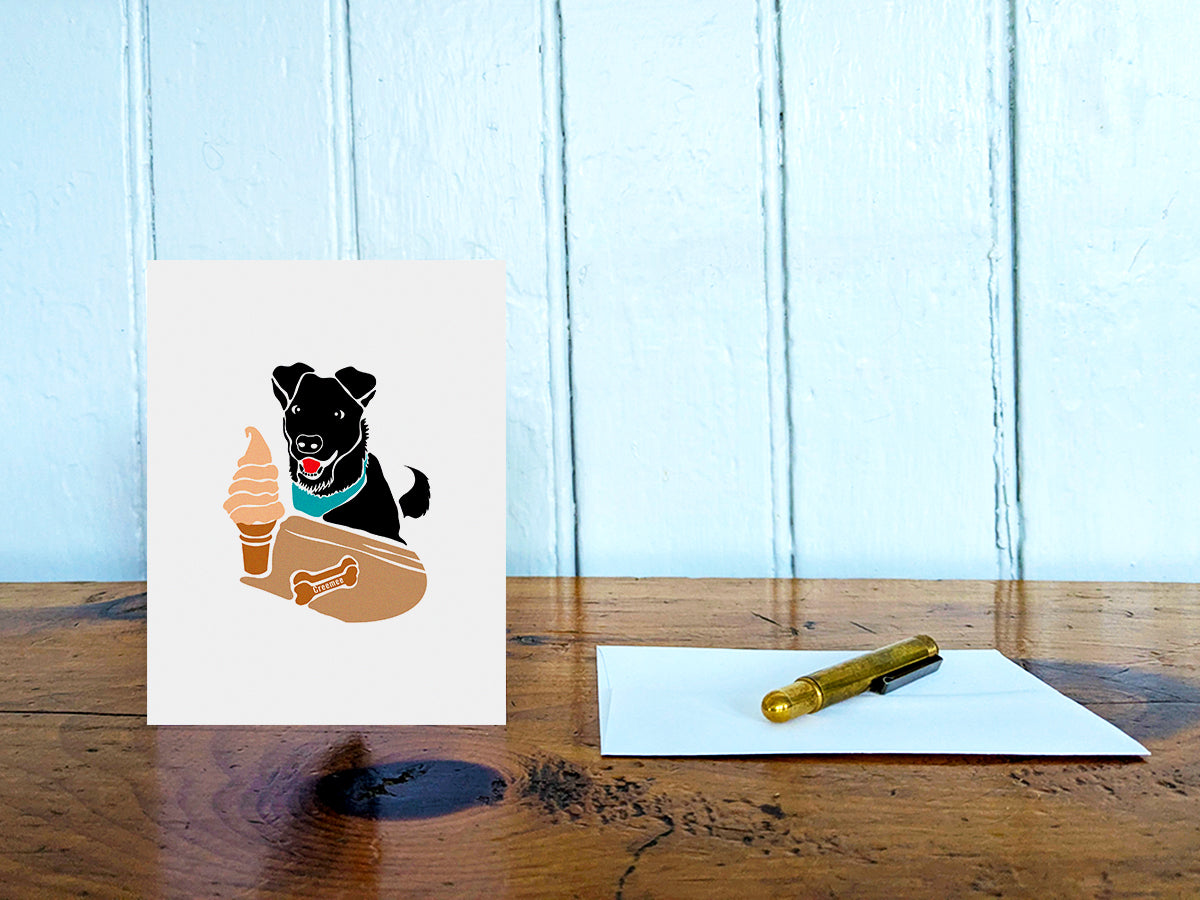 Greeting card with image of black dog looking at a creemee ice cream on a table with gold pen and envelope.