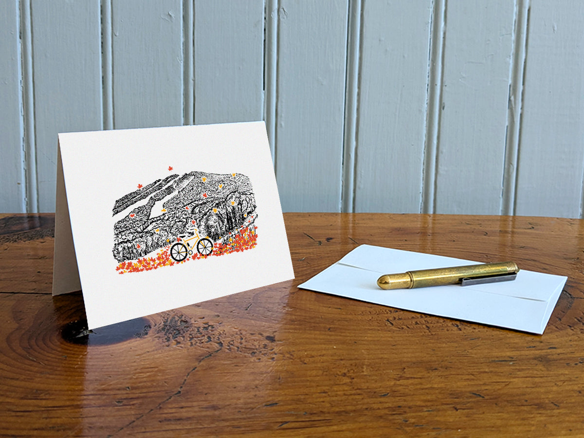 Greeting card with image of a mountain bike surrounded by brightly colored leaves with a  mountain in the background on a table with gold pen and envelope.