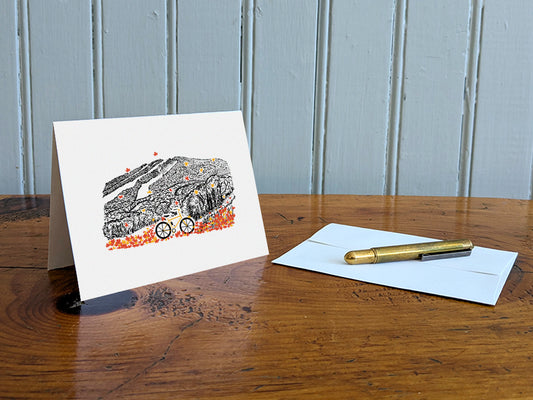 Greeting card with image of a mountain bike surrounded by brightly colored leaves with a  mountain in the background on a table with gold pen and envelope.