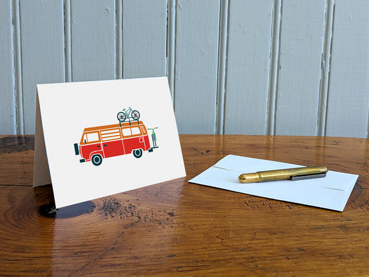 Greeting card with a red and orange camper van carrying two bikes on a table with gold pen and envelope