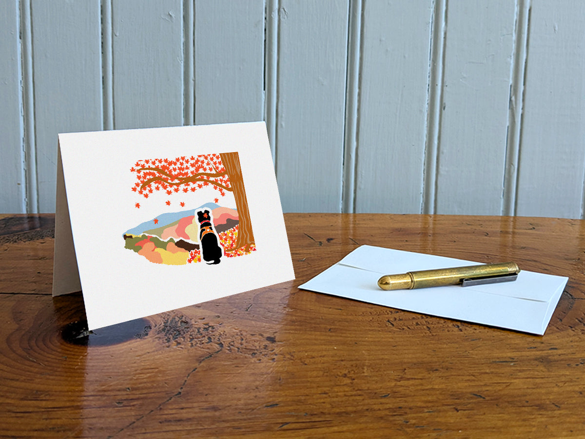 Greeting card with image of black dog underneath a maple tree with red fall foliage and mountains in the background on a table with gold pen and envelope.