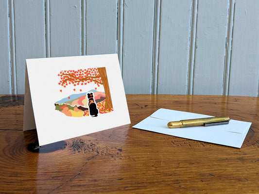 Greeting card with image of black dog underneath a maple tree with red fall foliage and mountains in the background on a table with gold pen and envelope.