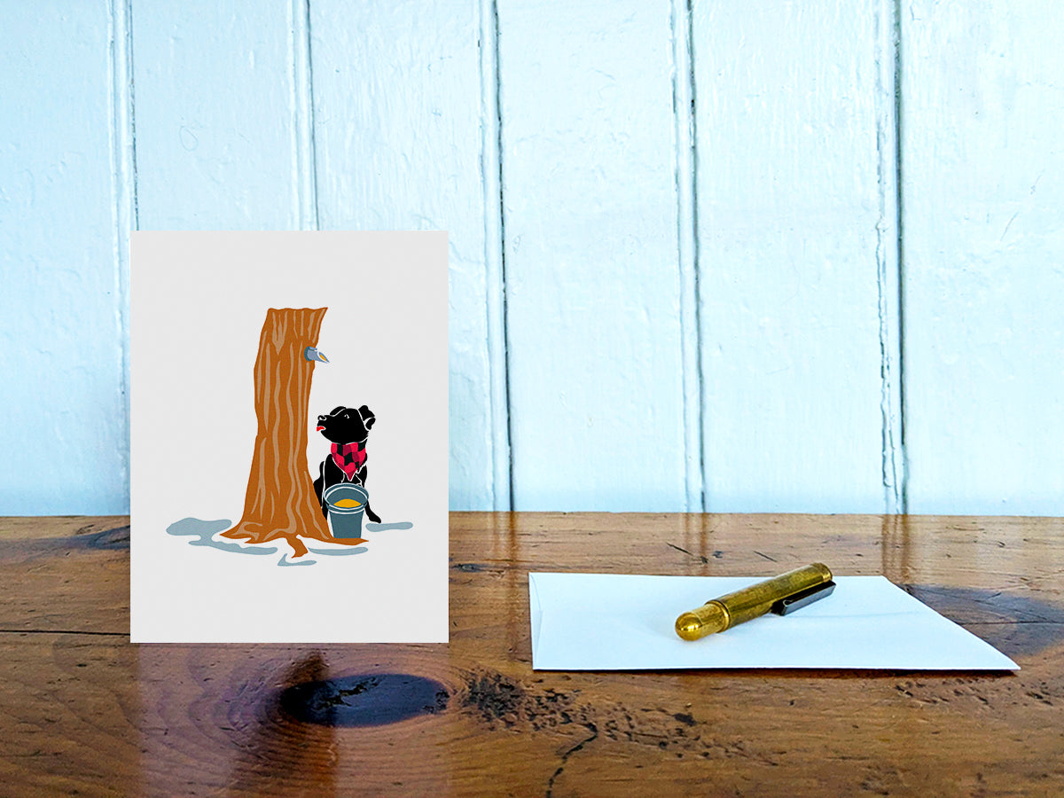 Greeting card with image of dog next to a maple syrup tap on a tree and bucket of maple syrup on a table with gold pen and envelope.