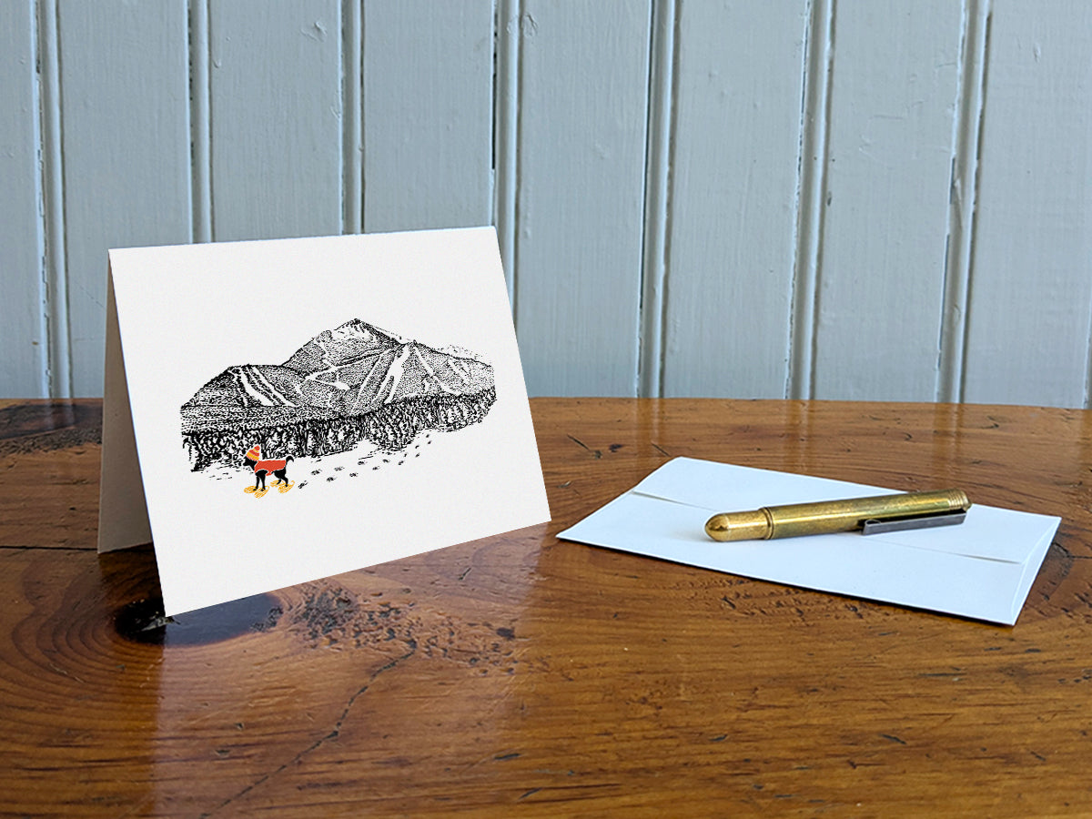 Greeting card with image of a mountain with ski trails and dog snowshoeing on a table with gold pen and envelope.