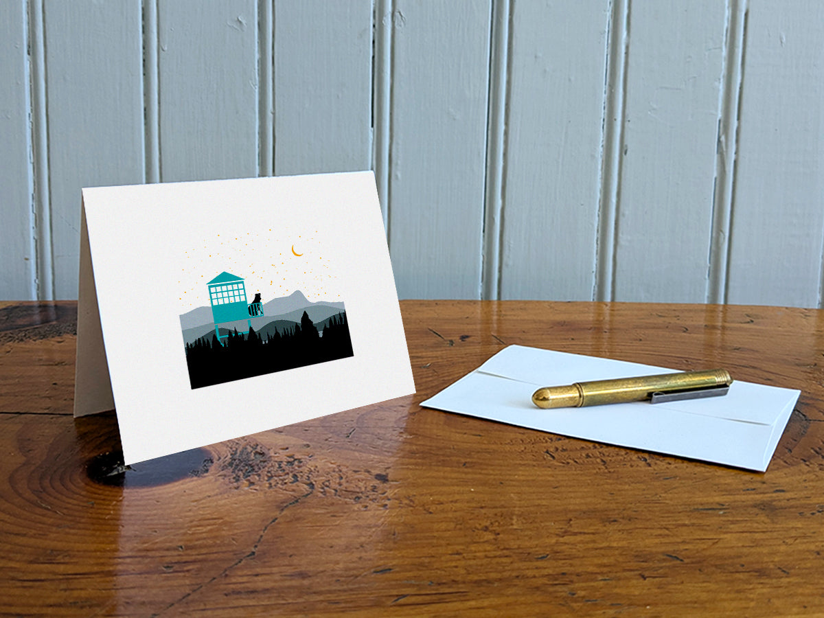 Greeting card of black dog sitting in a fire tower overlooking the mountains and stars on a table with gold pen and envelope.