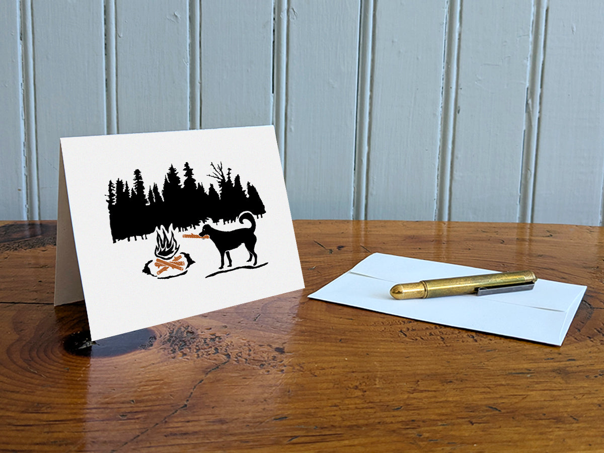 Greeting card of black dog putting a log in a campfire with forest background on a table with gold pen and envelope.