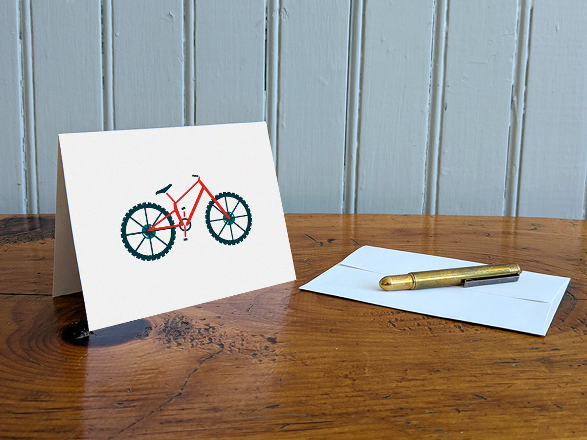 Greeting card with a red mountain bike with basket on a table with gold pen and envelope