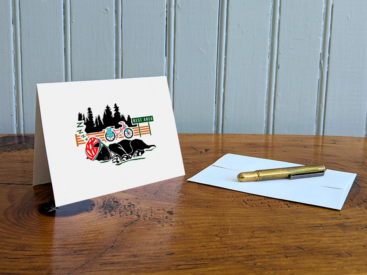 Greeting card of black dog with a red helmet sleeping with a bike and sign that reads Rest Area in the background on a table with gold pen and envelope