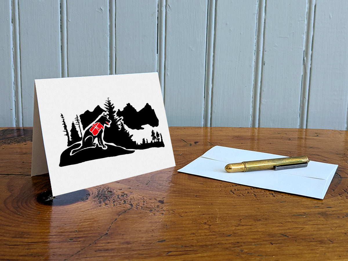 Greeting card with black dog with red backpack in the mountains on table with gold pen and envelope.