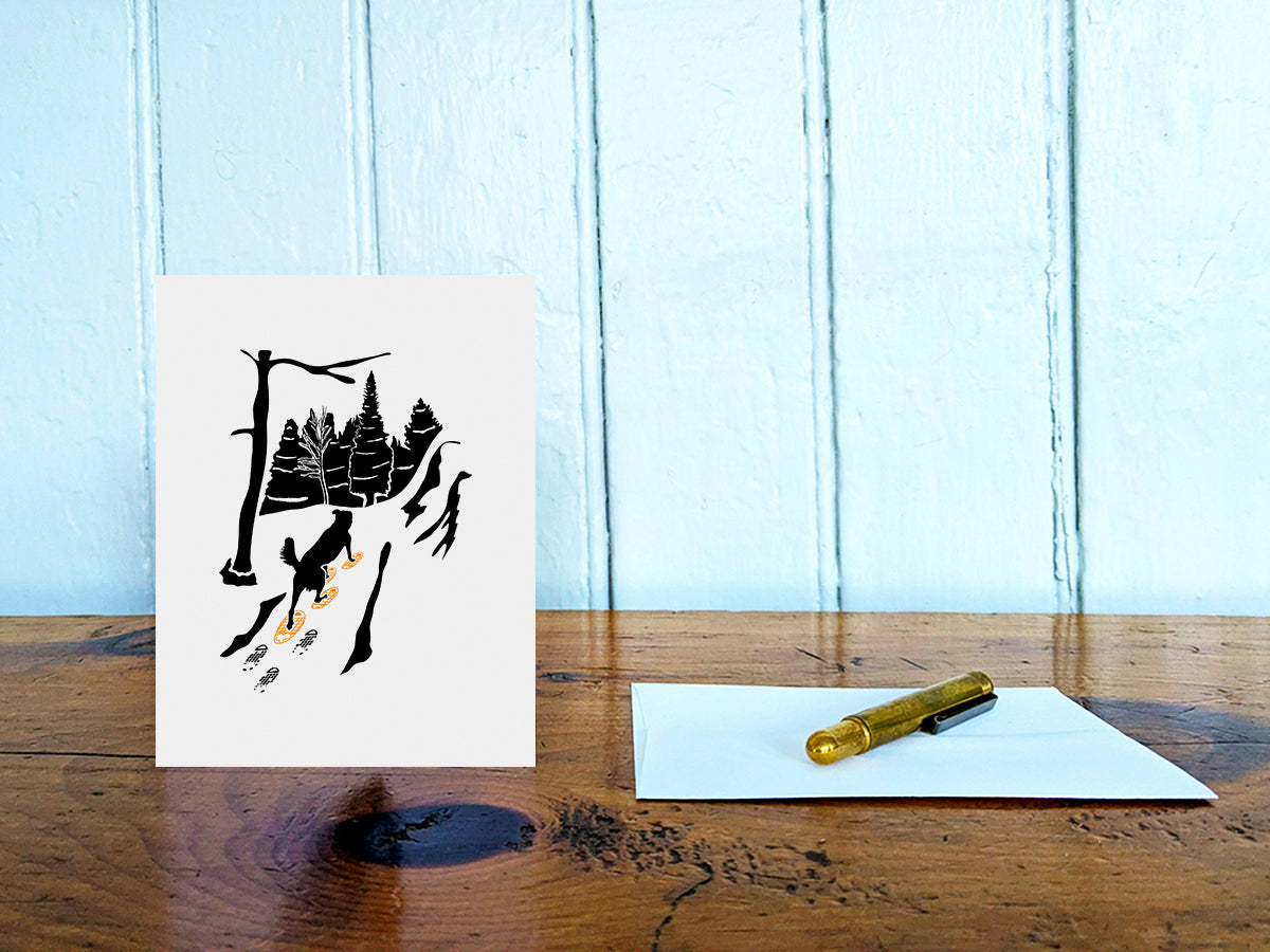 Greeting card with image of dog walking through forest wearing yellow snowshoes on table with gold pen and envelope.