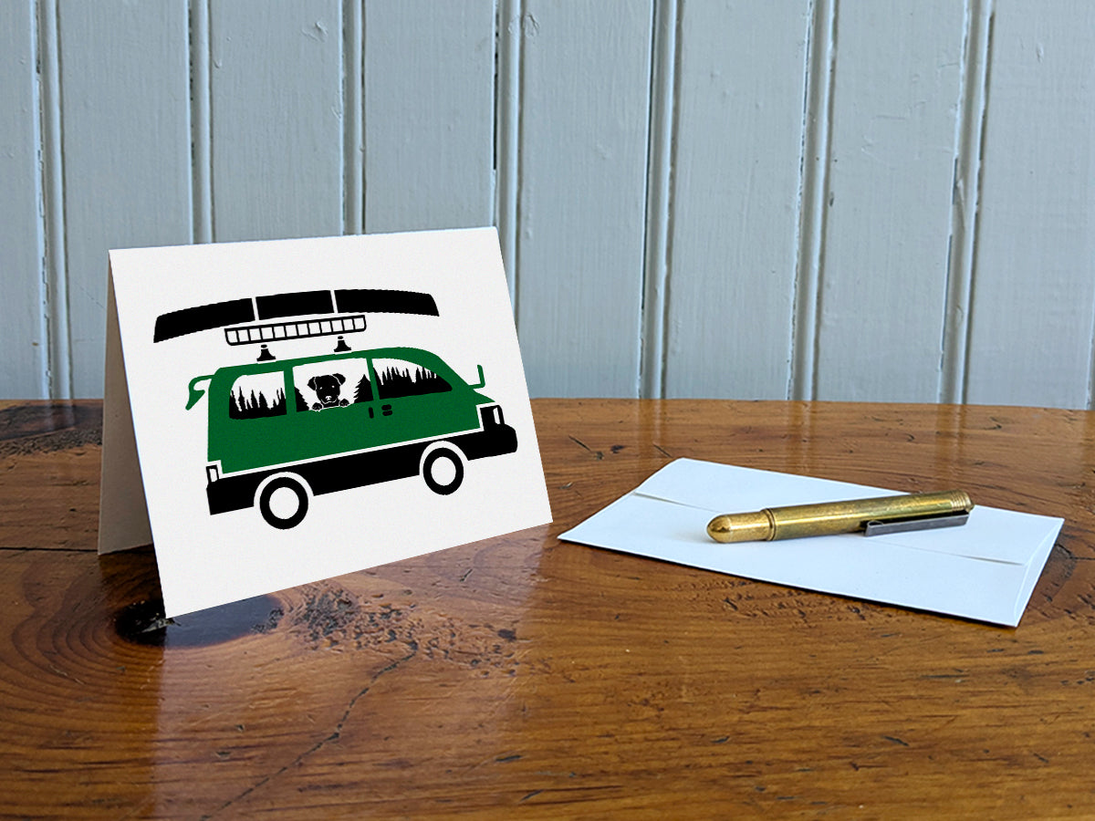 Greeting card with image of dog riding in a camper van with canoe on top on a table with gold pen and envelope.