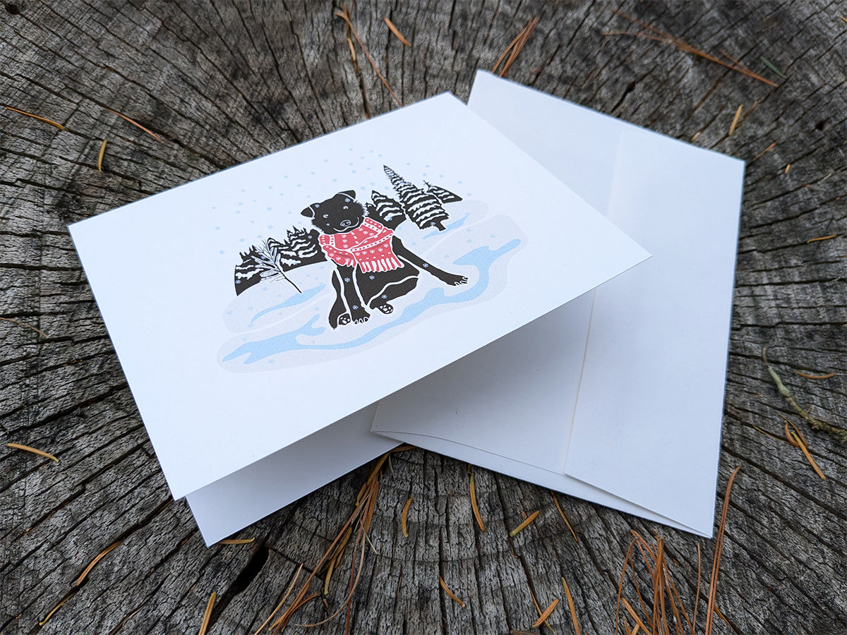 Greeting card with image of black dog with red scarf among a snowy winter landscape.