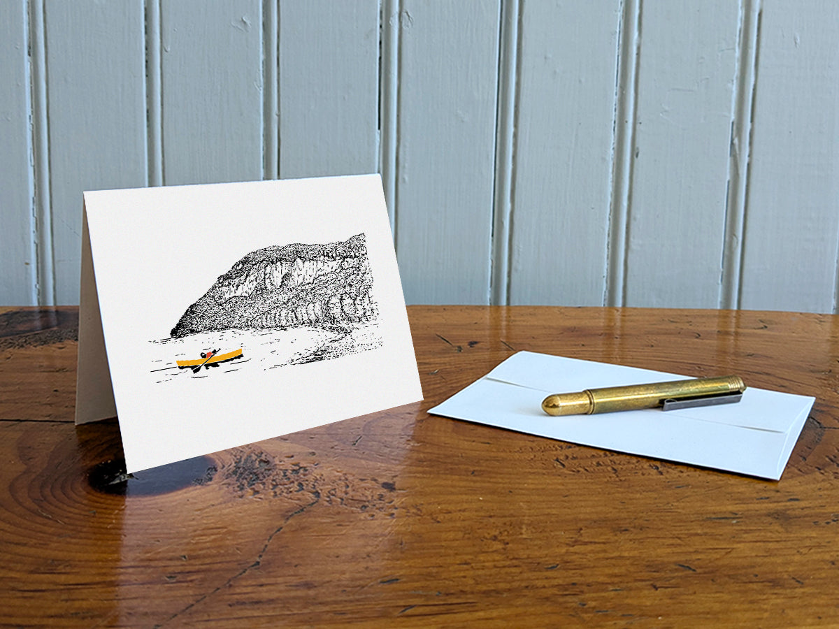 Greeting card featuring a dog paddling a canoe with mountain in the background on a table with gold pen and envelope.