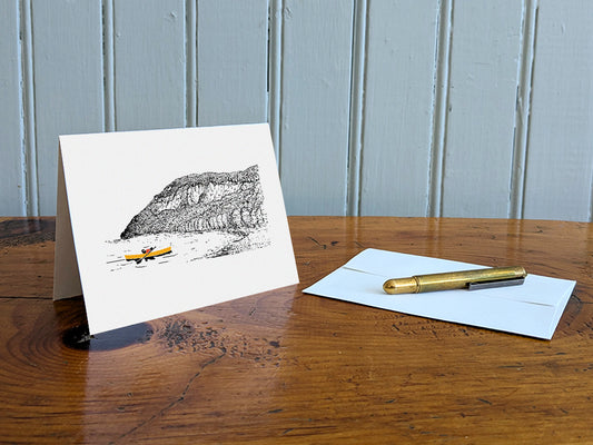 Greeting card featuring a dog paddling a canoe with mountain in the background on a table with gold pen and envelope.
