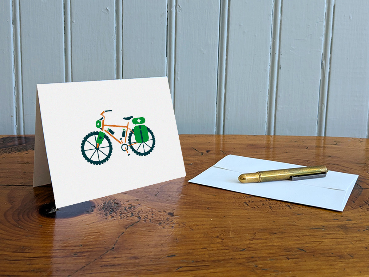 Greeting card with an orange bike with green travel bags on a table with gold pen and envelope