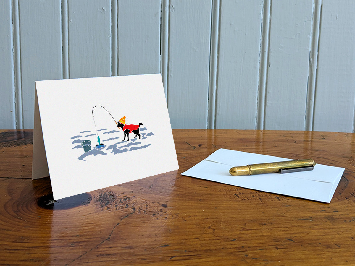 Greeting card with image of a dog with a winter hat and sweater ice fishing on a table with gold pen and envelope.