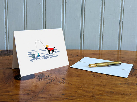 Greeting card with image of a dog with a winter hat and sweater ice fishing on a table with gold pen and envelope.