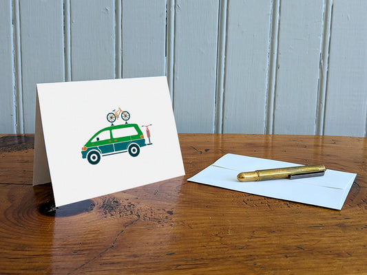 Greeting card with a blue and green van carrying two bikes on a table with gold pen and envelope
