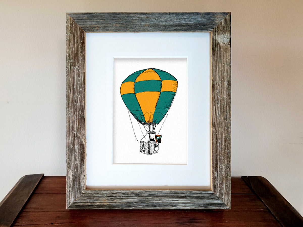 Framed illustration with a green and yellow hot air balloon with a black dog inside the basket.