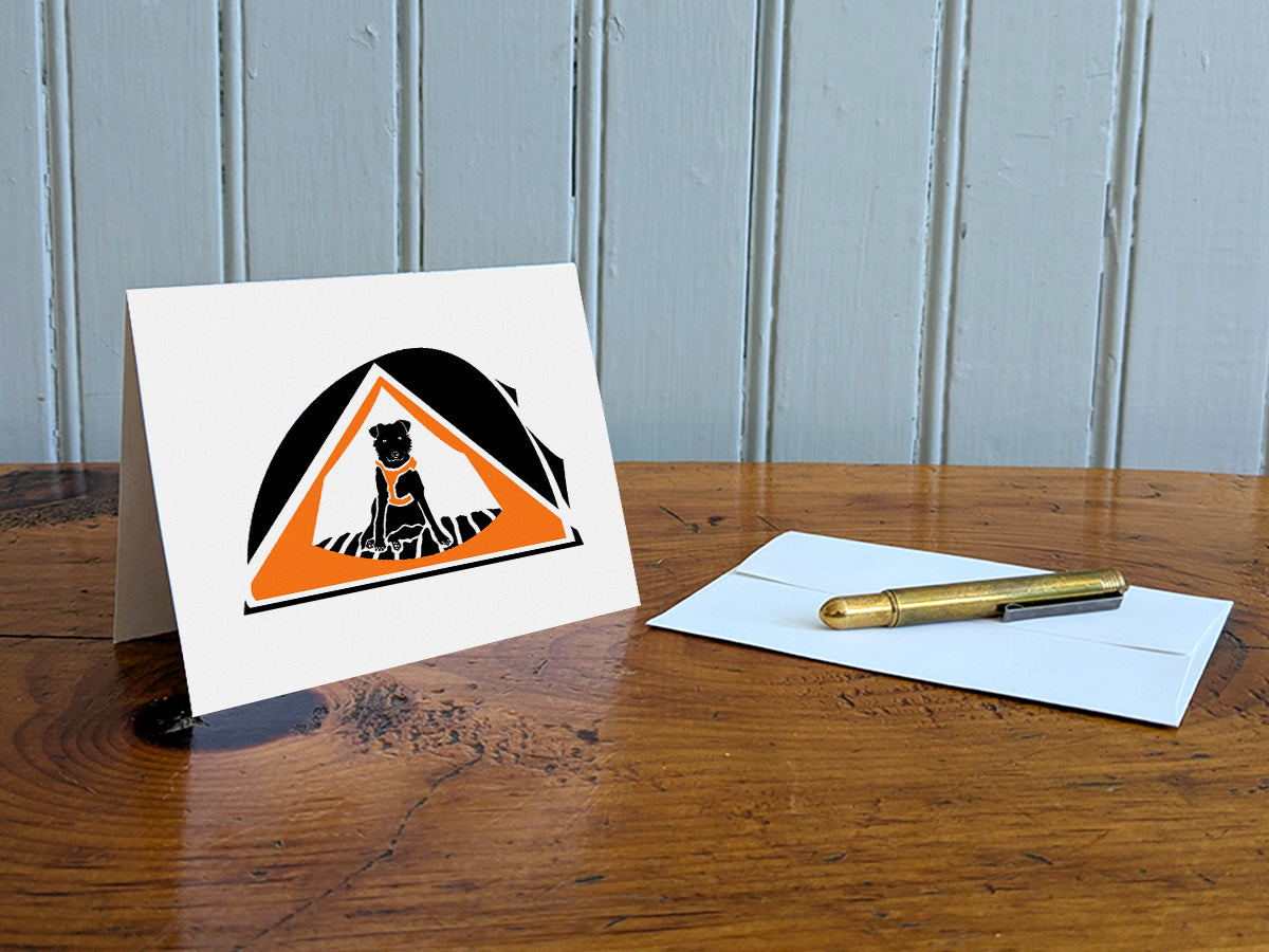 Greeting card with black dog with orange harness sitting in an orange camping tent on a table with gold pen and envelope.