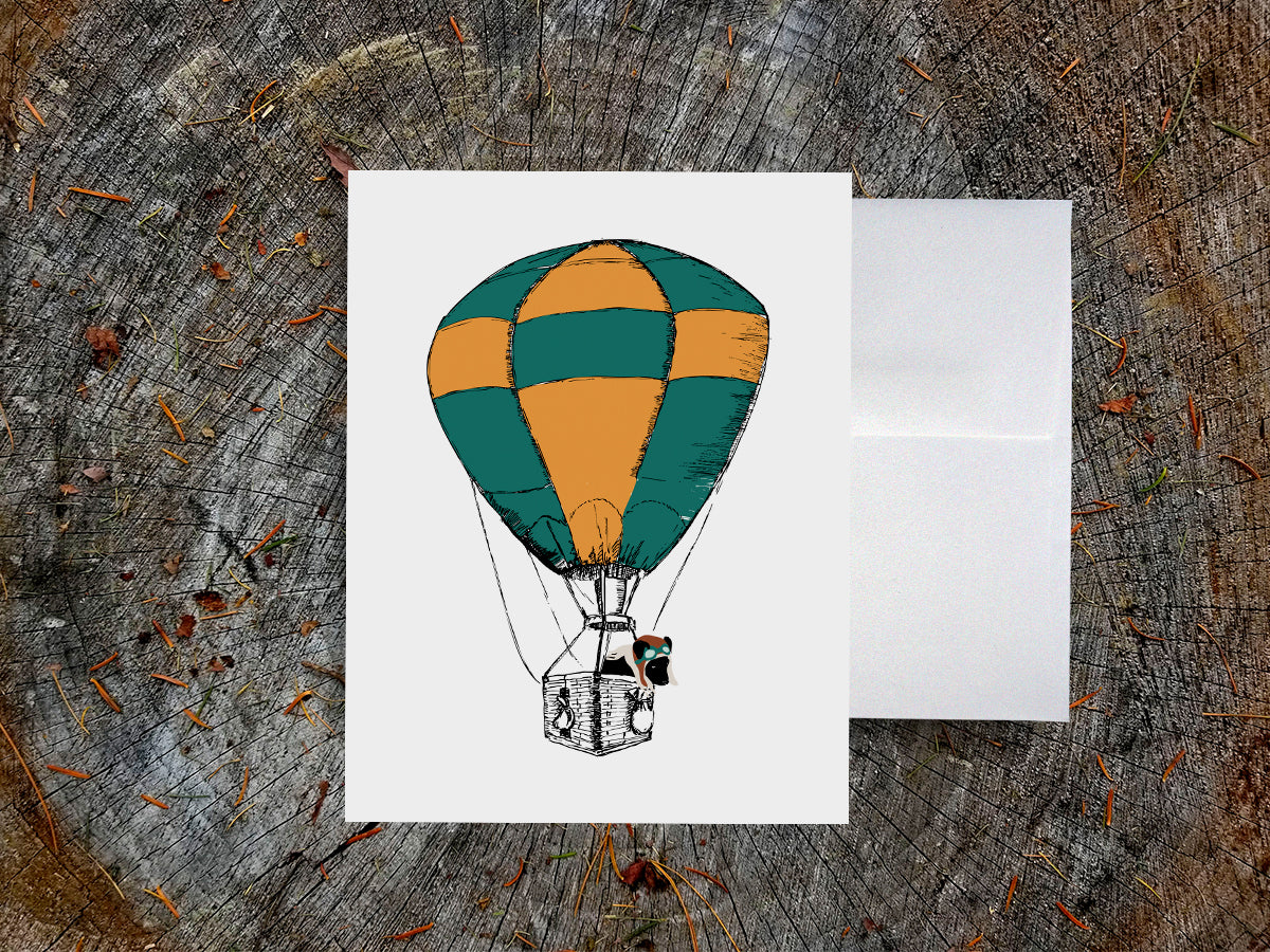 Greeting card with illustration of green and yellow hot air balloon with a black dog inside the basket on a wooden surface