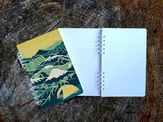 Notebook with illustration of a yellow tent among a background of blue and green mountains.
