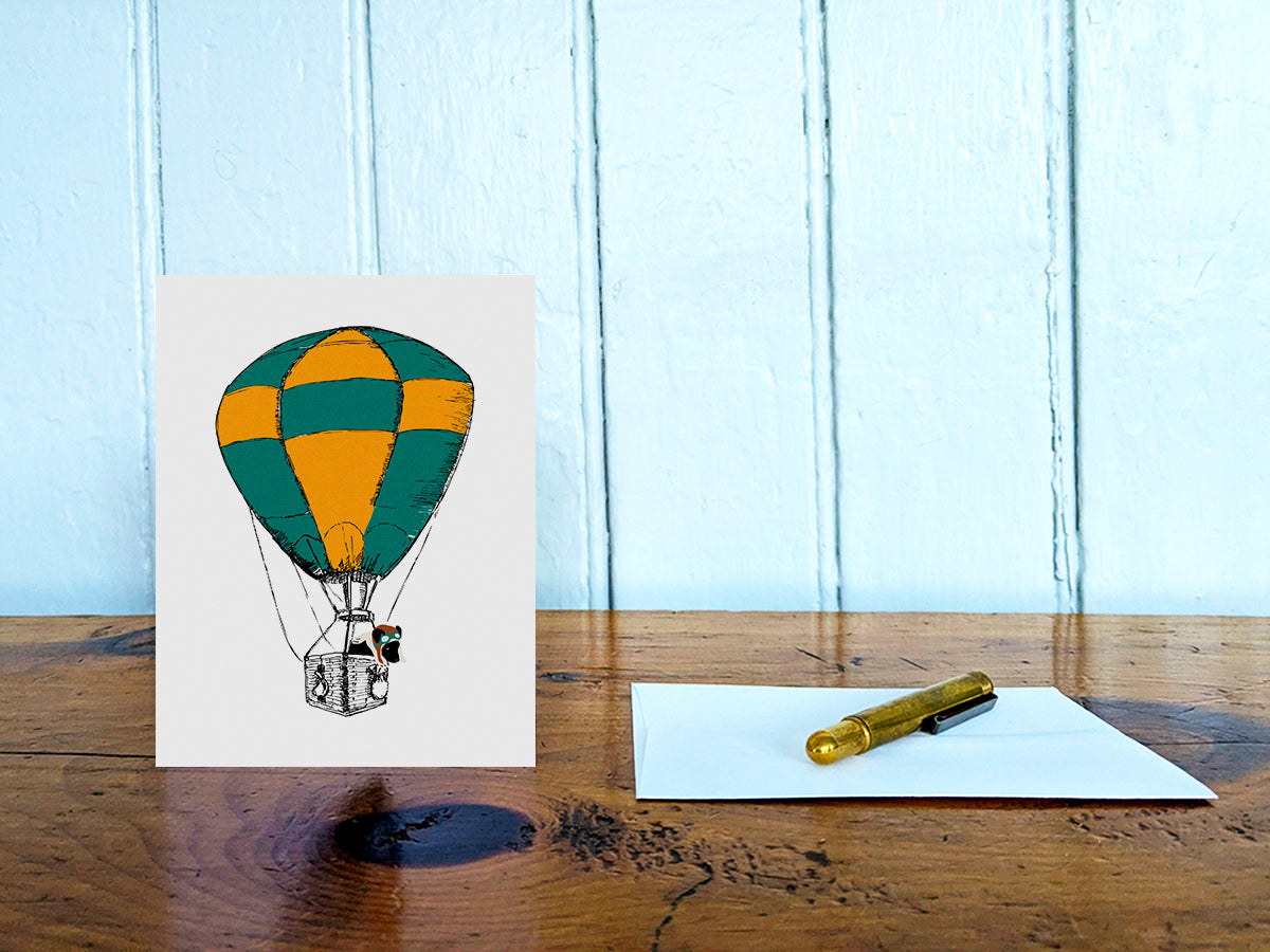 Greeting card with illustration of green and yellow hot air balloon with a black dog inside the basket on a wooden table with envelope and gold pen.