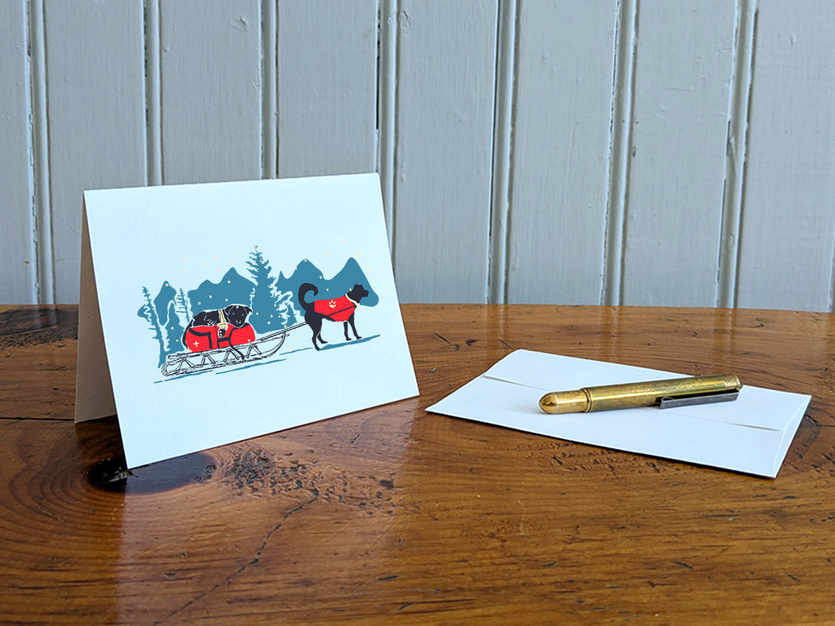 Greeting card with dog in a red jacket pulling a sled with another dog in a wintry landscape, on a wooden surface next to a white envelope and gold pen.
